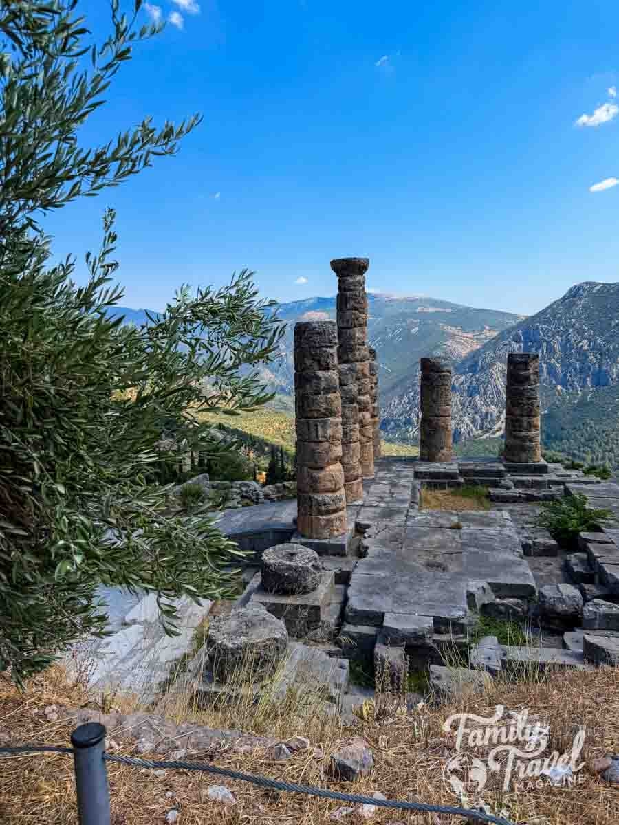 Ancient stone columns stand on a rocky terrace overlooking a mountainous landscape under a clear blue sky in Delphi