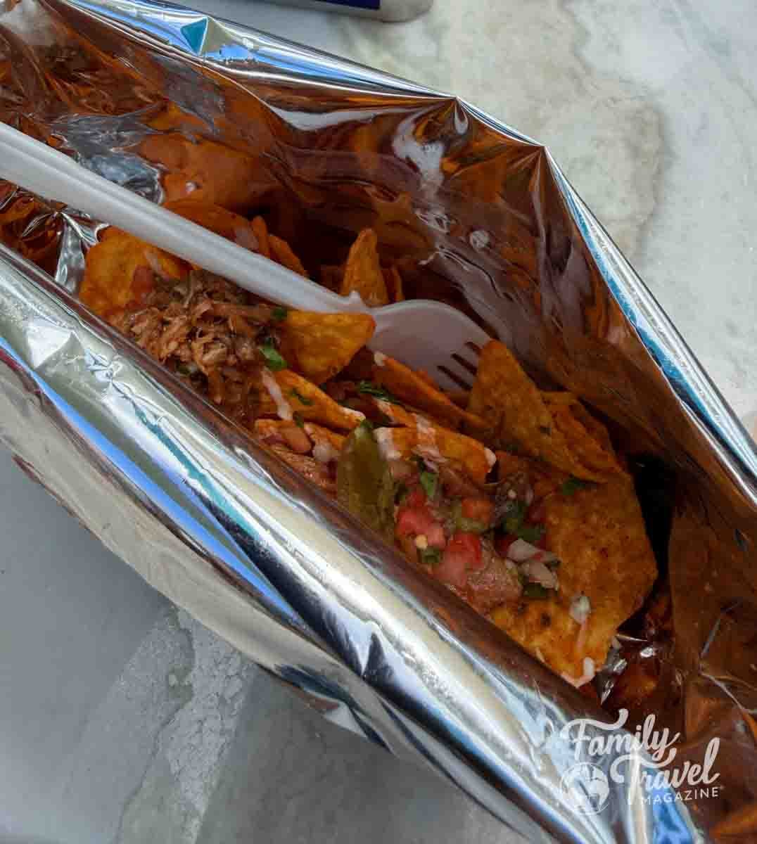 A plastic fork rests inside a metallic foil bag filled with tortilla chips topped with meat, salsa, and peppers.
