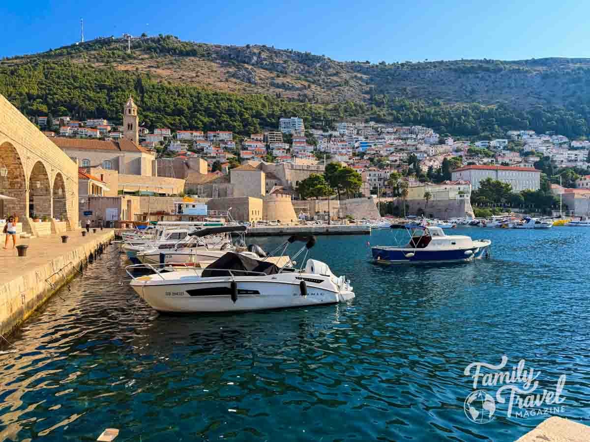 Small boats are docked in a harbor in front of a coastal town with stone buildings and a hillside in Dubrovnik. 