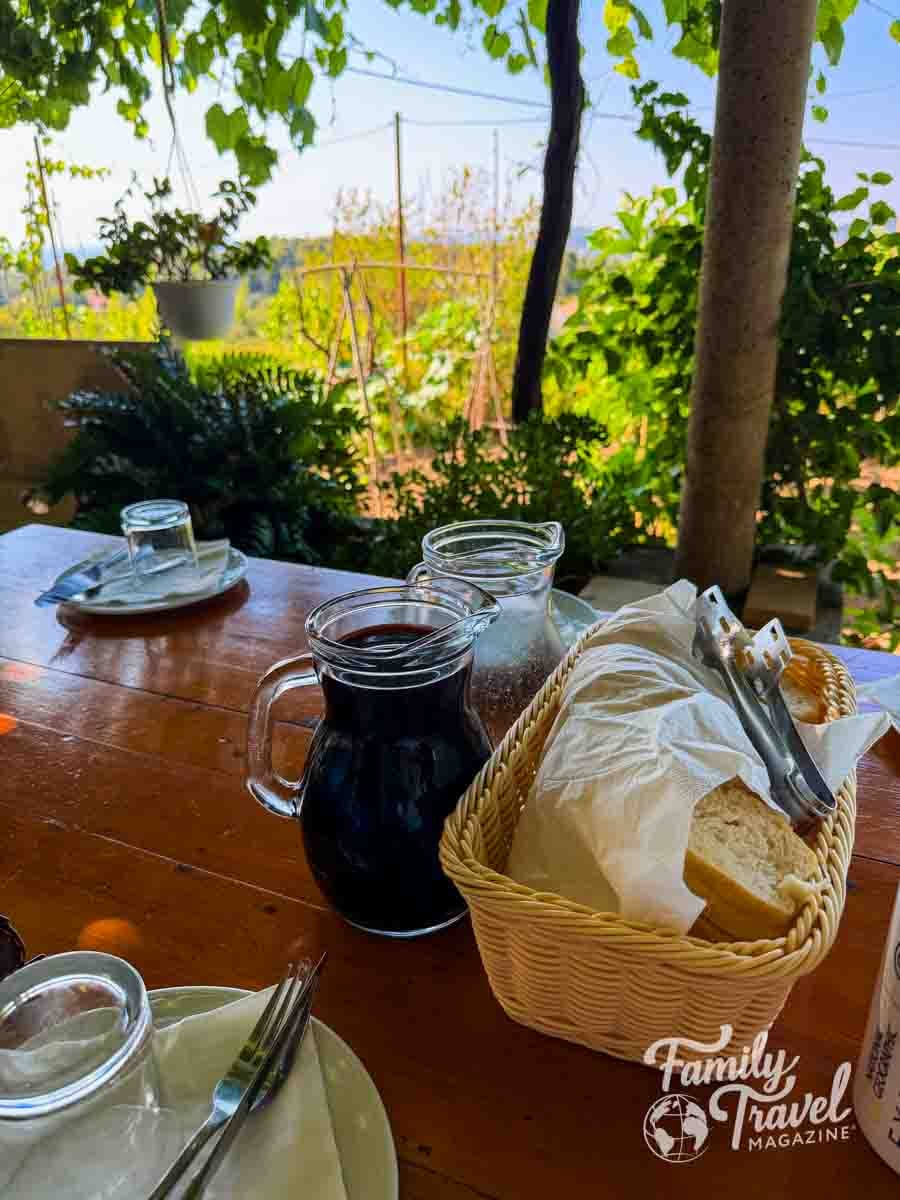 A wooden table is set with a basket of bread, a pitcher of red liquid, and glasses, overlooking a sunny outdoor garden.