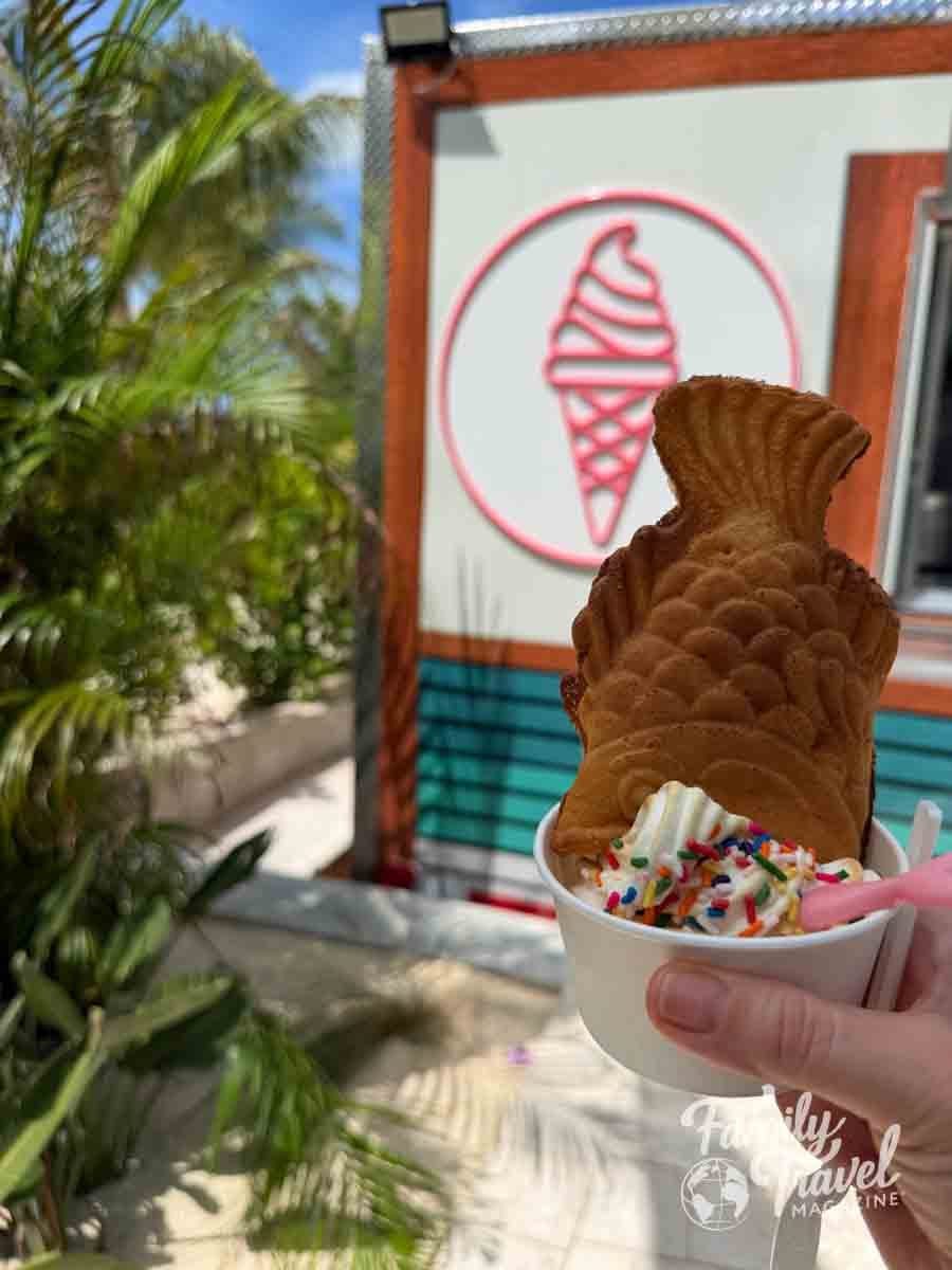 A hand holds a cup of soft serve ice cream topped with sprinkles and a fish-shaped waffle in front of a food stand.