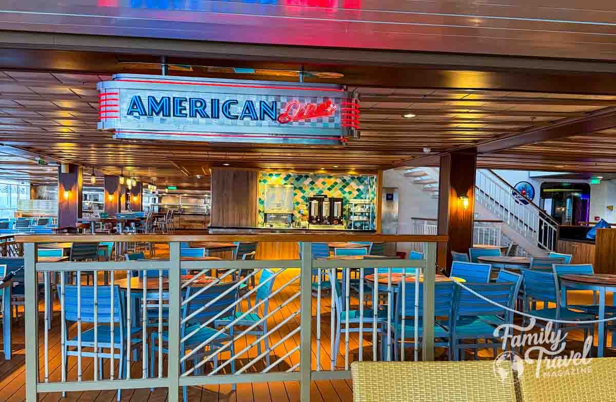 A restaurant dining area features a retro-style sign reading AMERICAN above tables and blue chairs on a wooden deck.
