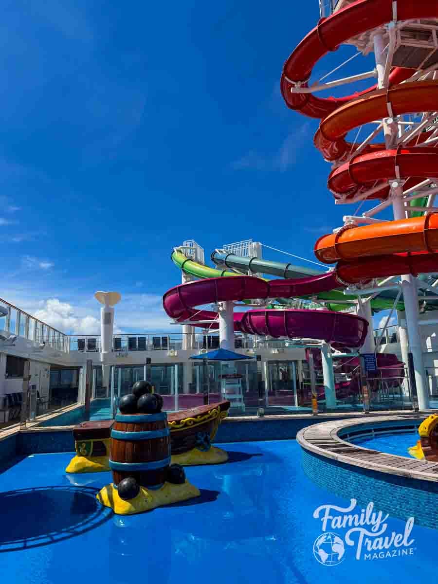 A multi-colored water slide structure towers over a shallow pool with a decorative barrel and ship-themed play feature.