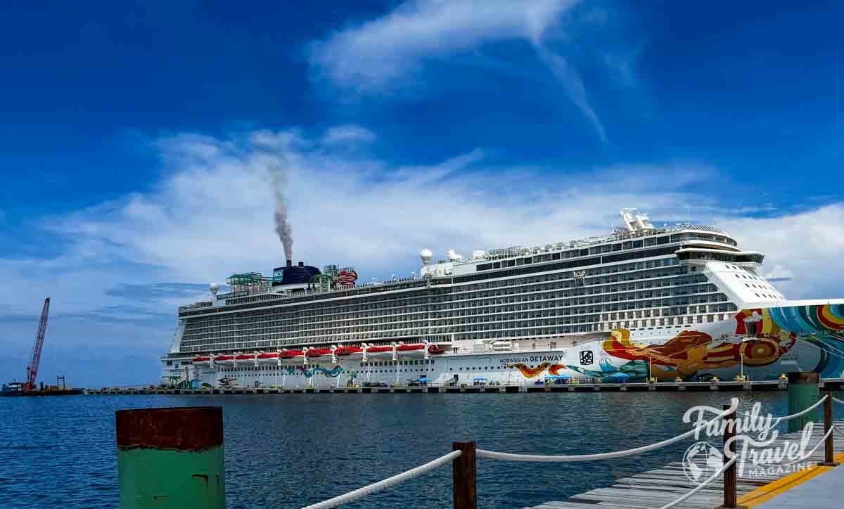 A large cruise ship is docked at a pier under a blue sky with light clouds.