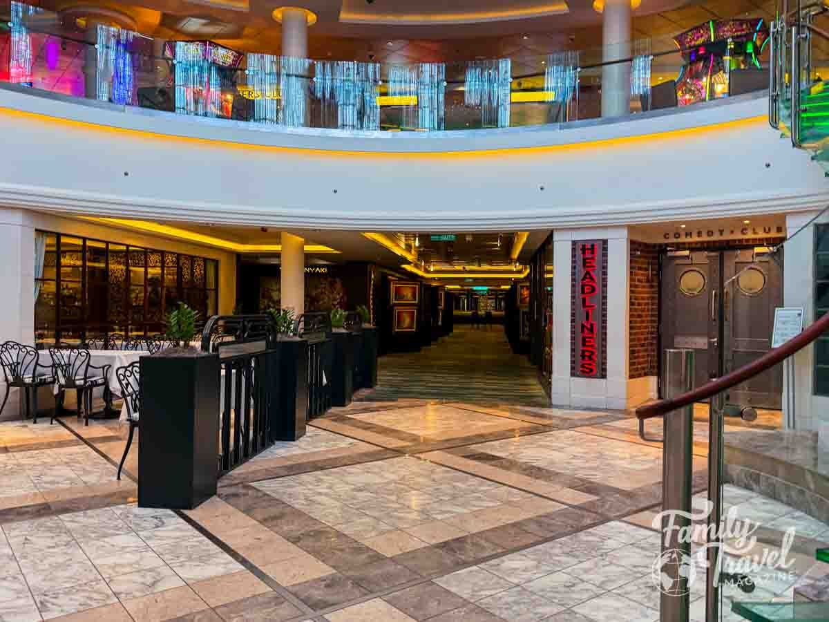 A multi-level indoor corridor features a dining area with tables and chairs, a comedy club entrance, and marble flooring.