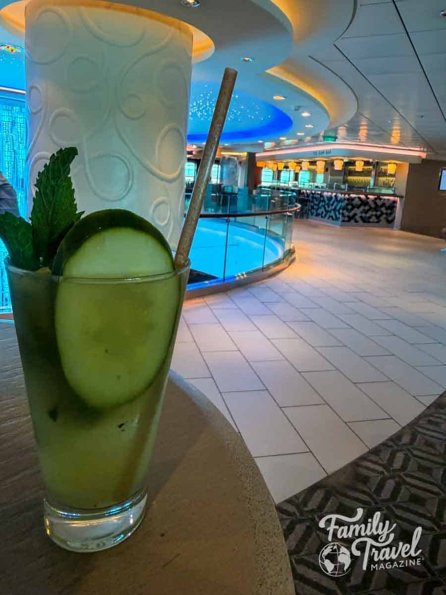 A glass with a cucumber slice and mint garnish sits on a table in front of a brightly lit indoor lounge area.