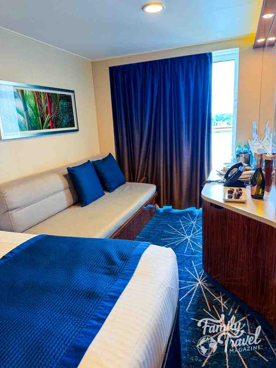 A hotel room features a bed with a blue runner, a sofa with blue pillows, and a desk with a bottle and glasses.