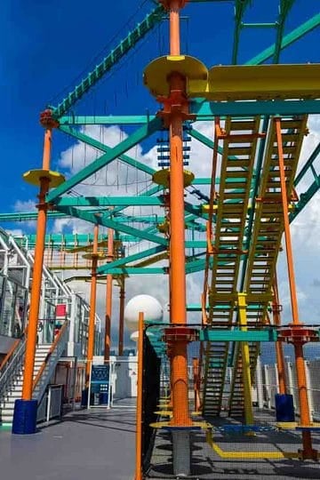 A tall, colorful ropes course structure stands on the open deck of a ship under a blue sky with clouds.