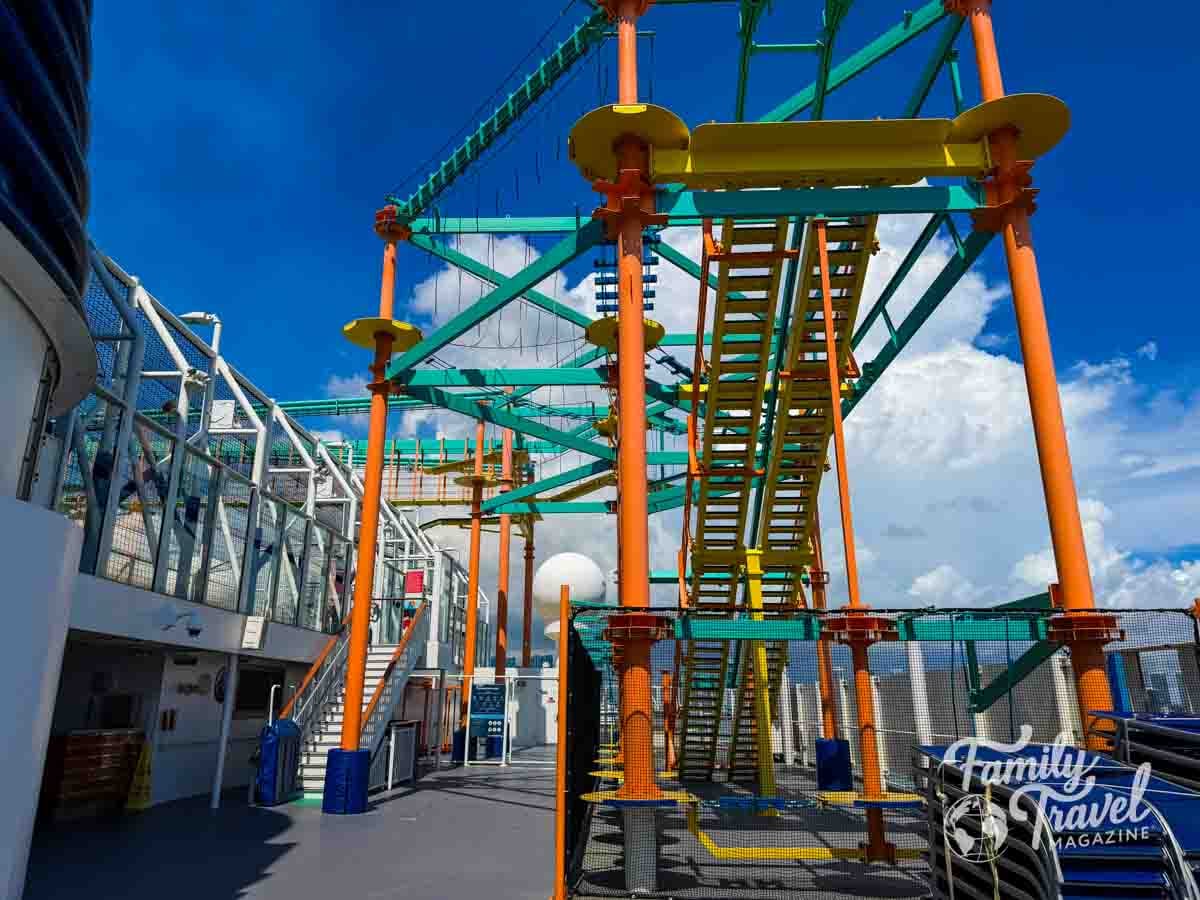 A tall, colorful ropes course structure stands on the open deck of a ship under a blue sky with clouds.