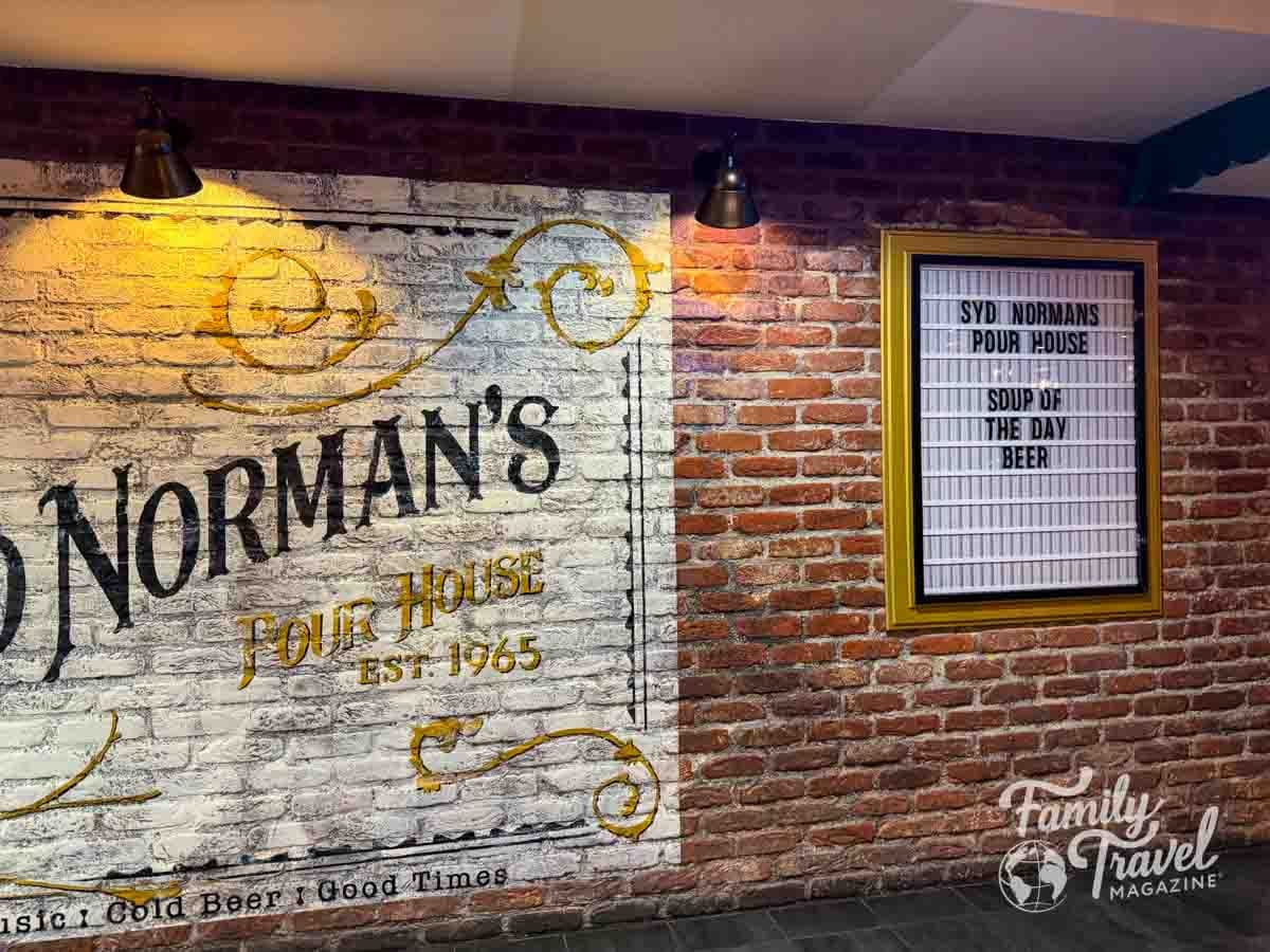 A brick wall features a painted sign for Normans Pour House and a framed menu board listing soup and beer.