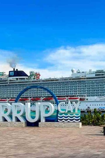 A large cruise ship is docked behind a sign that reads Great Stirrup Cay on a paved waterfront area under a blue sky.