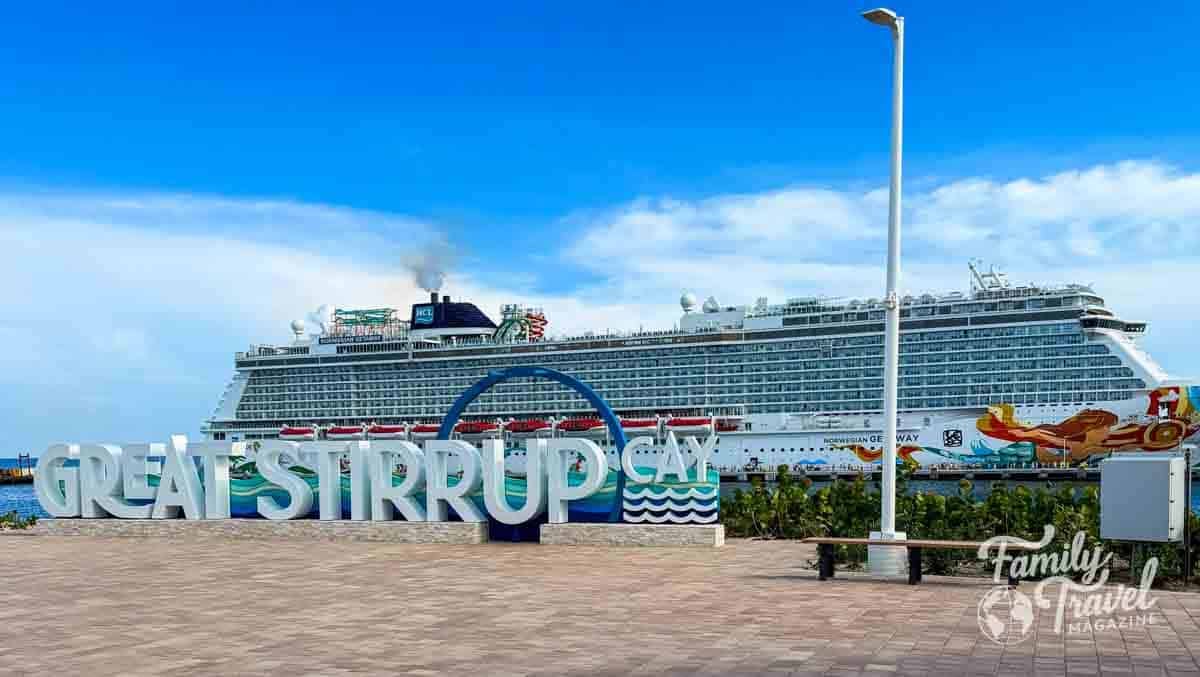 A large Norwegian cruise ship is docked behind a sign that reads Great Stirrup Cay on a paved waterfront area under a blue sky.