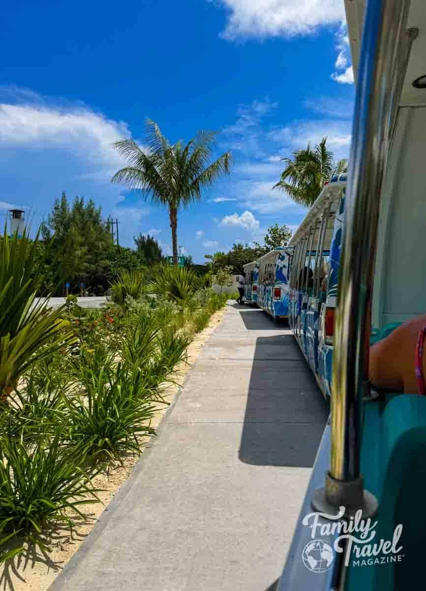 A multi-car tram travels along a concrete path lined with tropical plants and palm trees under a blue sky at Great Stirrup Cay.