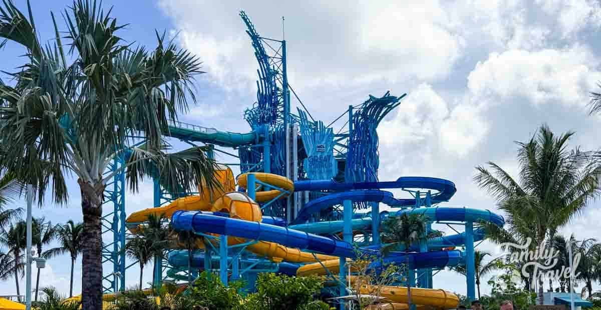 A large water park structure features multiple winding blue and yellow slides surrounded by palm trees under a cloudy sky at Norwegian Cruise Line's Great Tides Waterpark