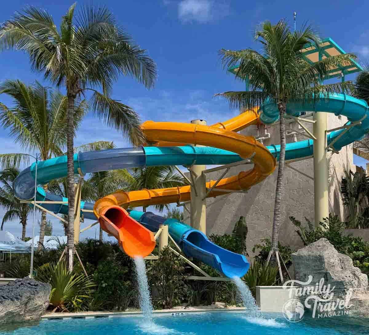 Two winding water slides in orange and blue empty into a swimming pool surrounded by palm trees under a clear blue sky.