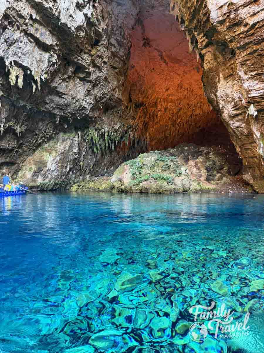 A small boat floats on clear blue water inside a large cavern with rocky walls and a partially open ceiling at Melissani Lake.