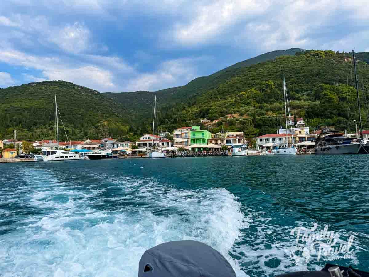A boat wake churns the blue water in front of a harbor lined with buildings and docked sailboats against green hills in Kefalonia Greece