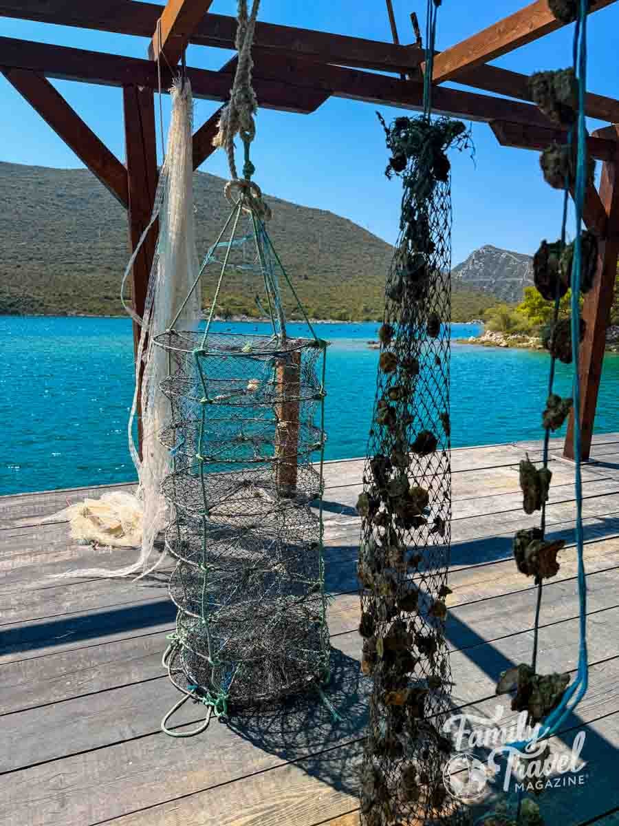 Fishing nets and a cylindrical wire trap hang from a wooden structure overlooking a blue bay and mountains.