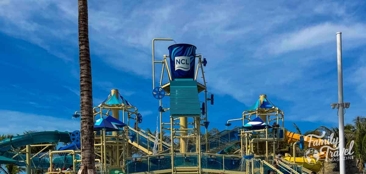 A water play structure features a large tipping bucket labeled NCL, slides, and platforms against a blue sky.