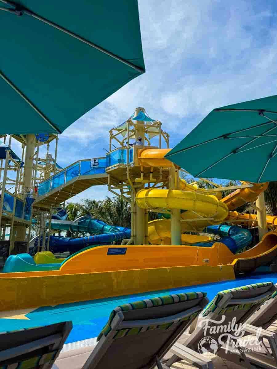A water park features yellow and blue slides under large teal umbrellas with lounge chairs in the foreground.