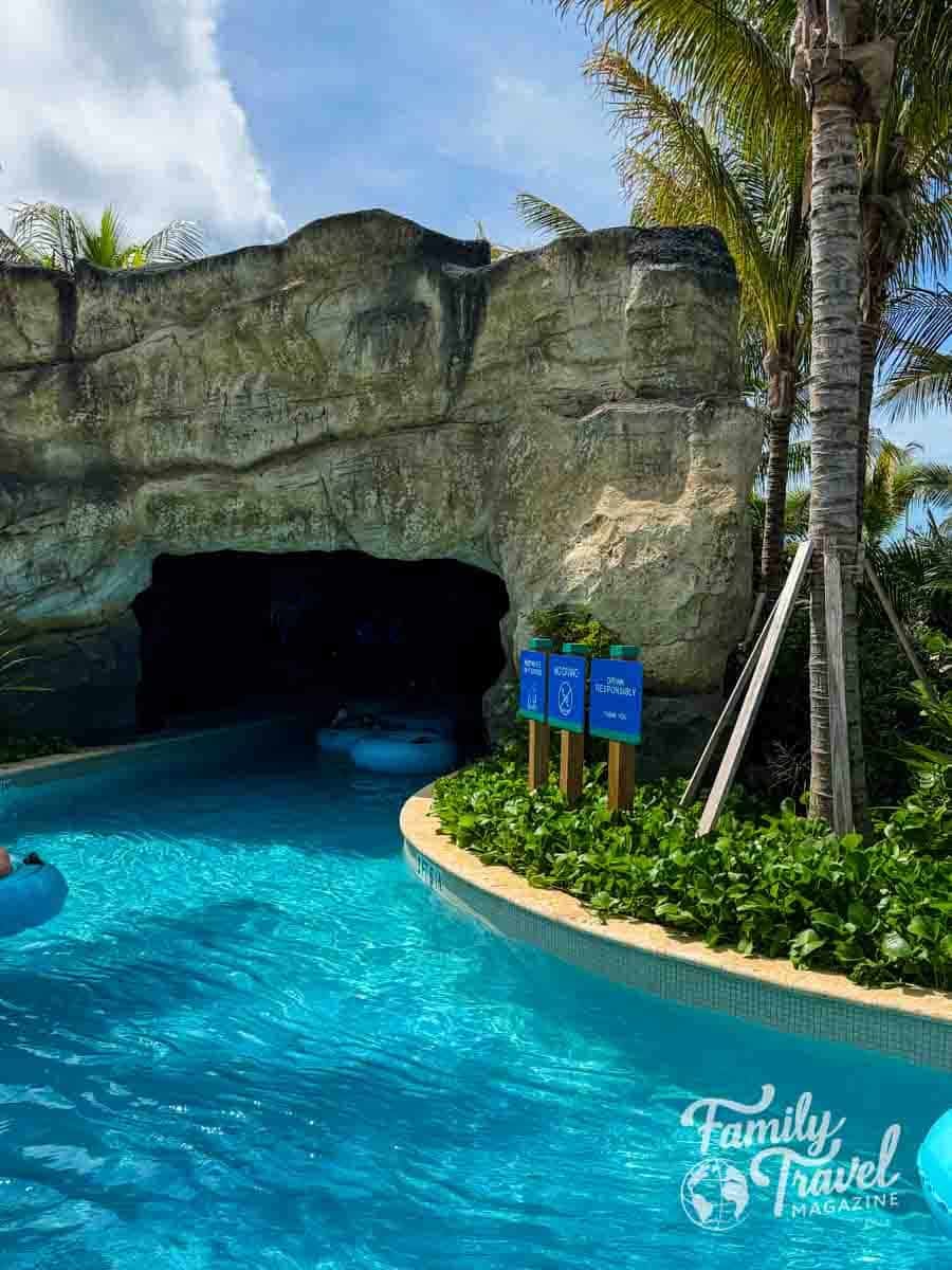 A lazy river flows into a dark cave entrance beside a rocky wall, palm trees, and small informational signs.