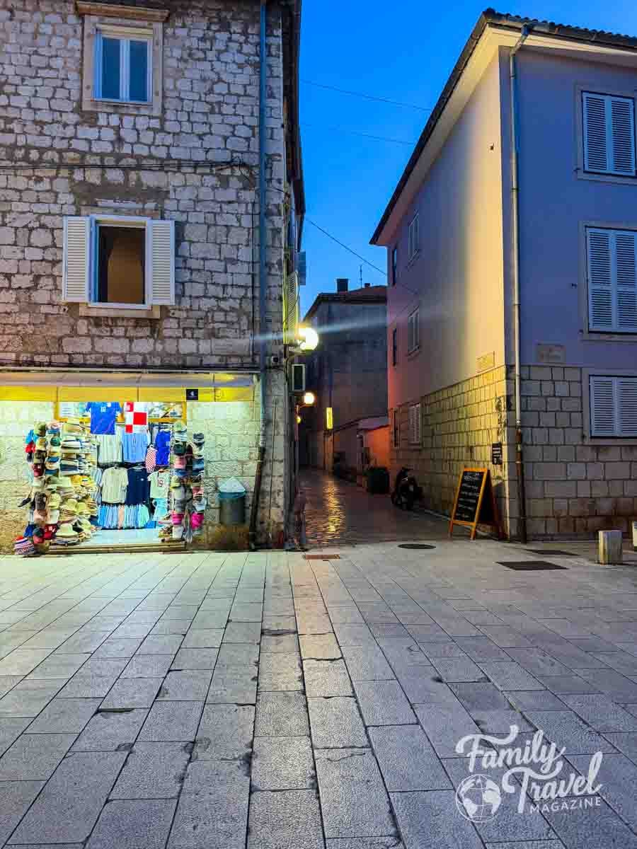 A narrow stone-paved street at dusk sits between two buildings, one featuring a brightly lit shop displaying clothing.