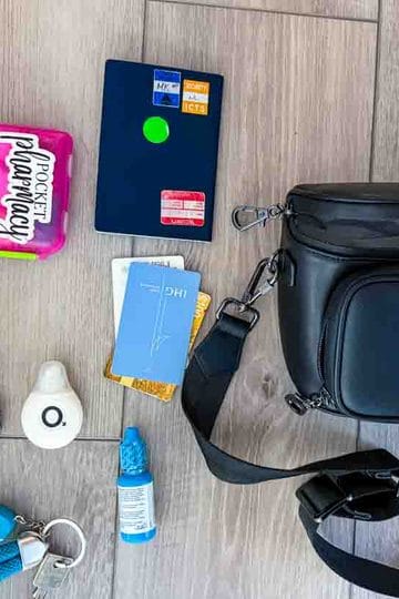 A black crossbody bag lies on a wooden floor next to a blue patterned pouch, a passport, cards, and various small items.
