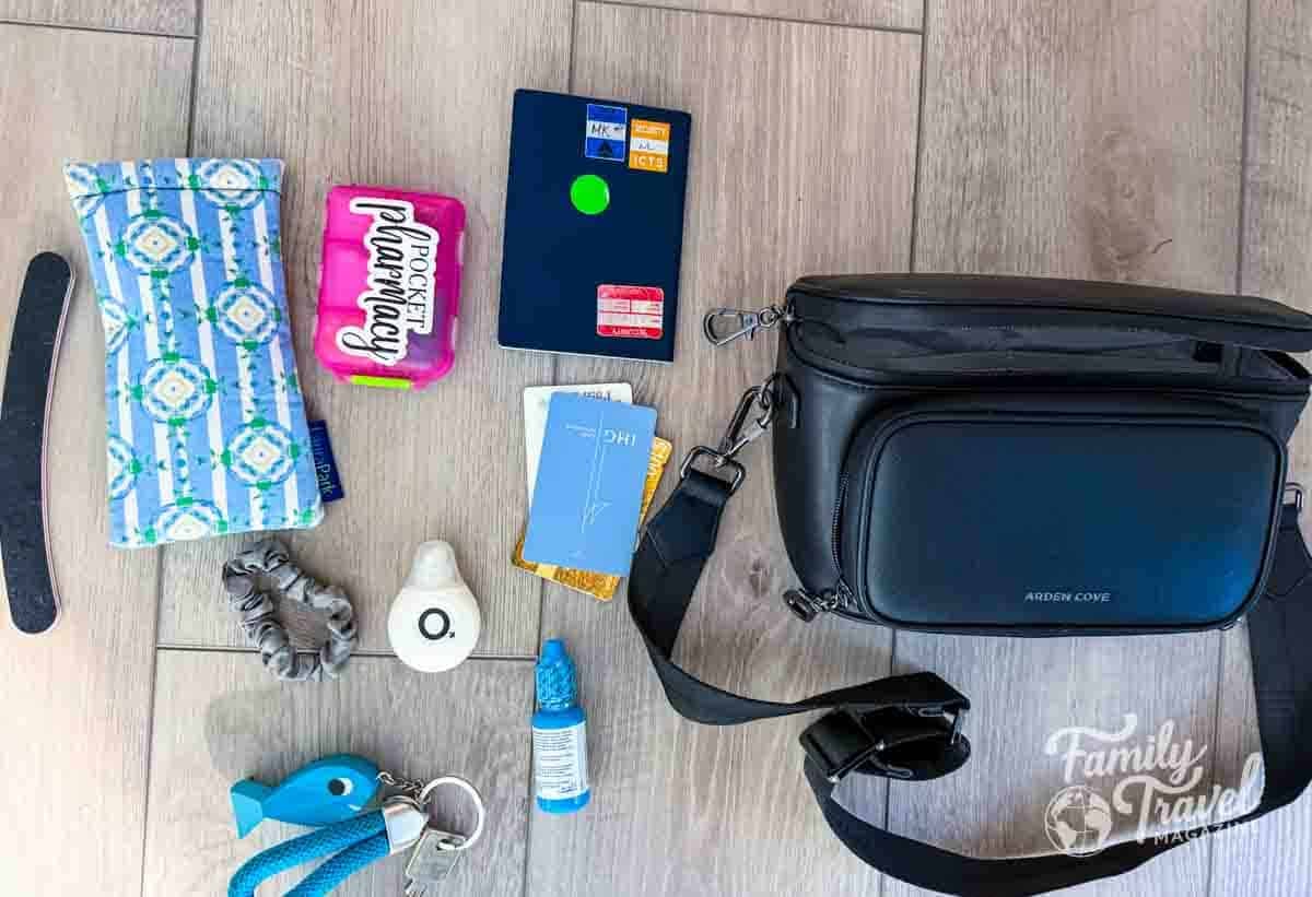 A black crossbody bag lies on a wooden floor next to a blue patterned pouch, a passport, cards, and various small items.