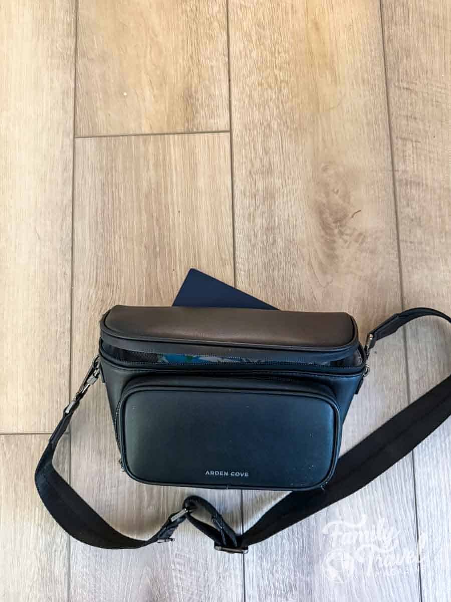 A black crossbody bag with a front pocket rests on a light wood-grain floor.