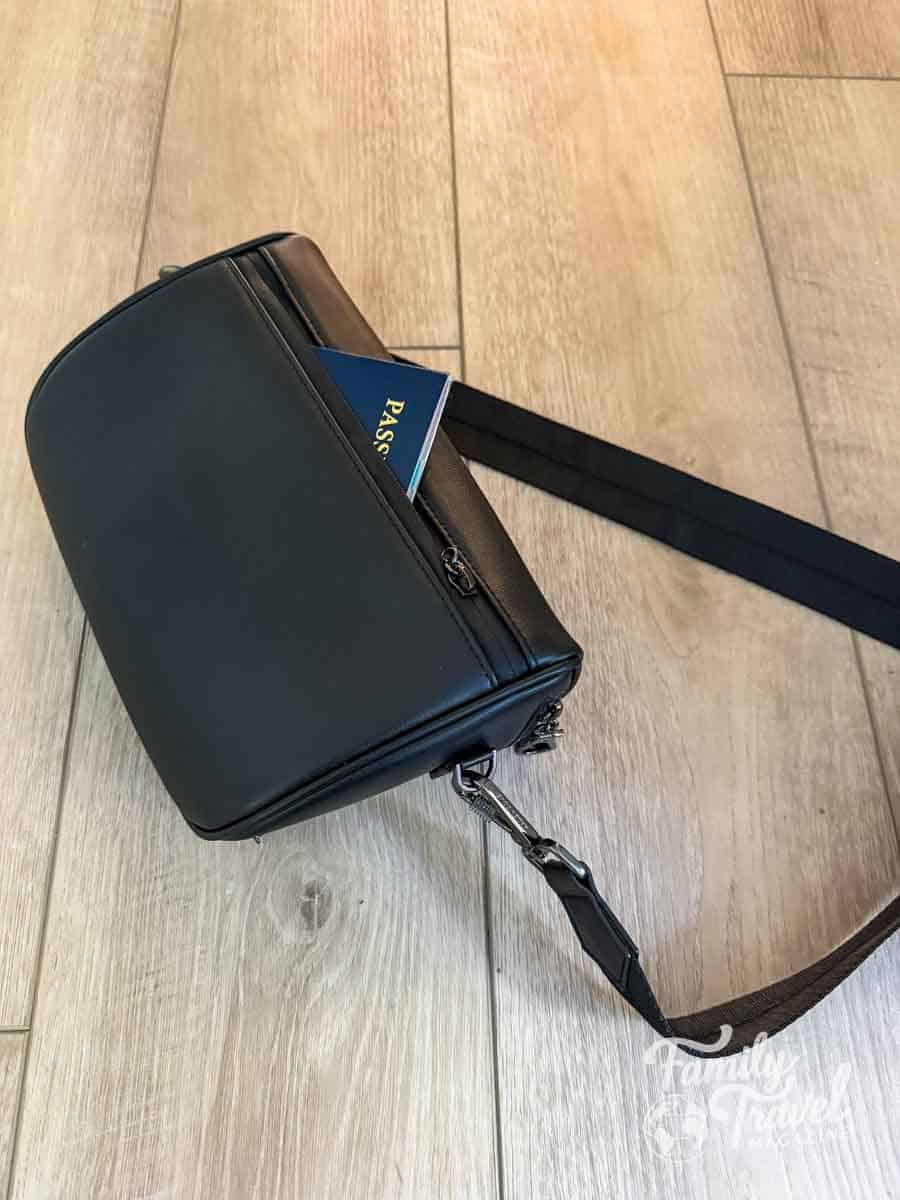 A black crossbody bag with a passport partially tucked into its outer pocket sits on a light wood-grain floor.